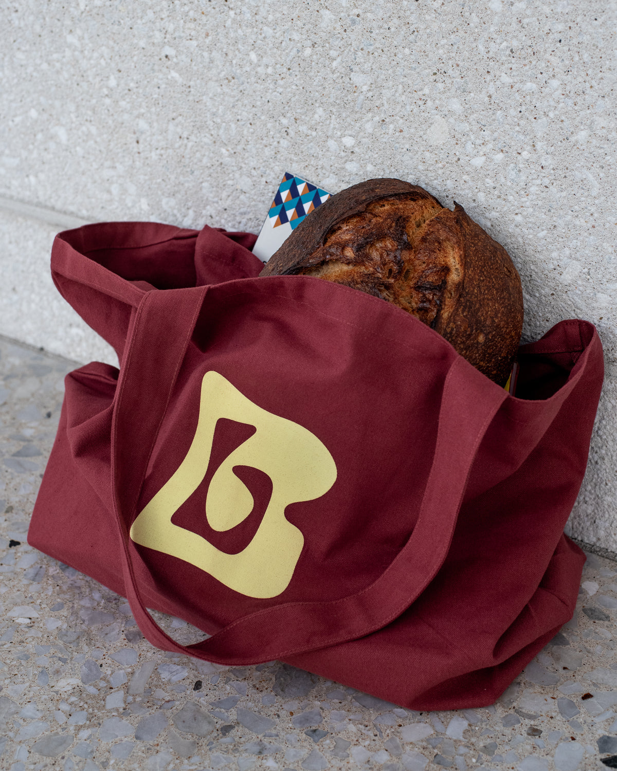 Burgundy tote bag with lemon yellow Berberlin logo sitting on grey marble. The large bag is holding bread and books.